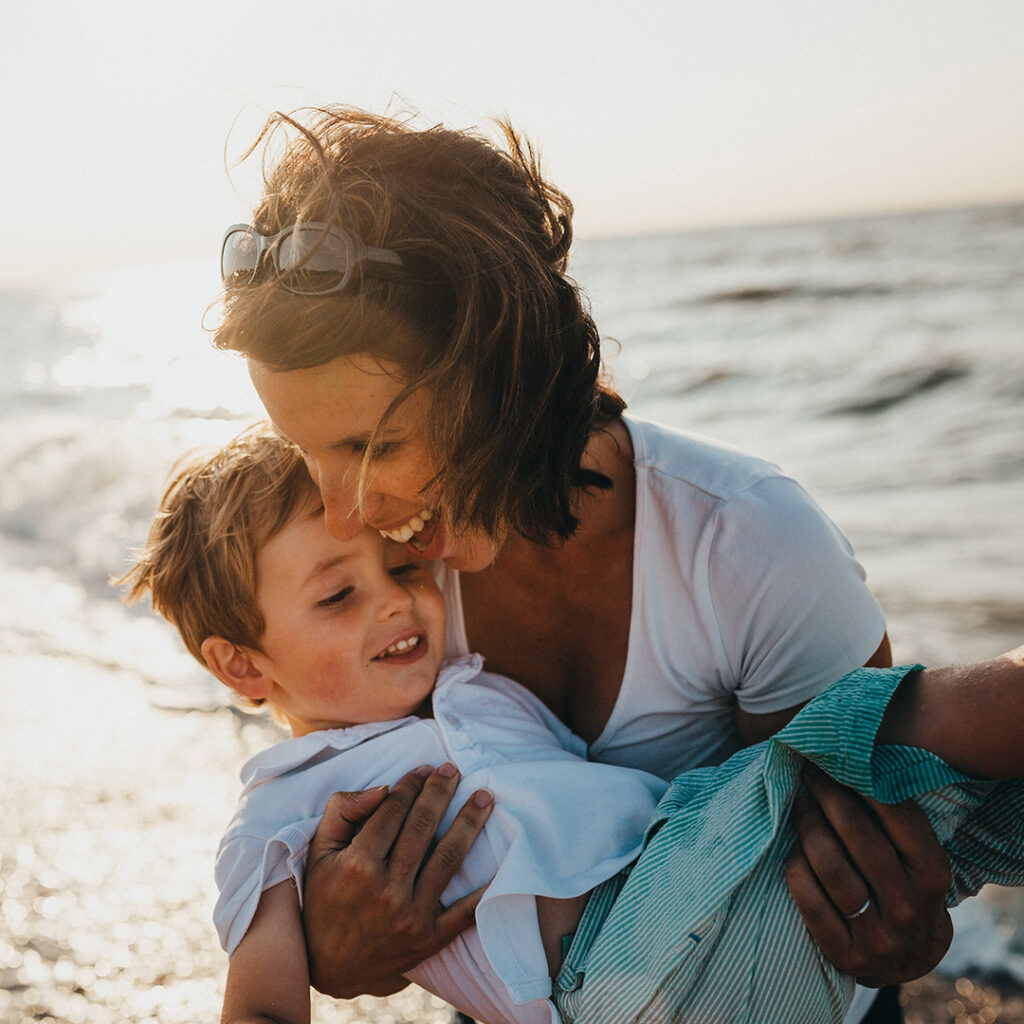 Woman joyfully holding her son at the beach, both smiling and looking healthy, symbolizing the positive impact of stem cell autoimmune care treatment on her well-being